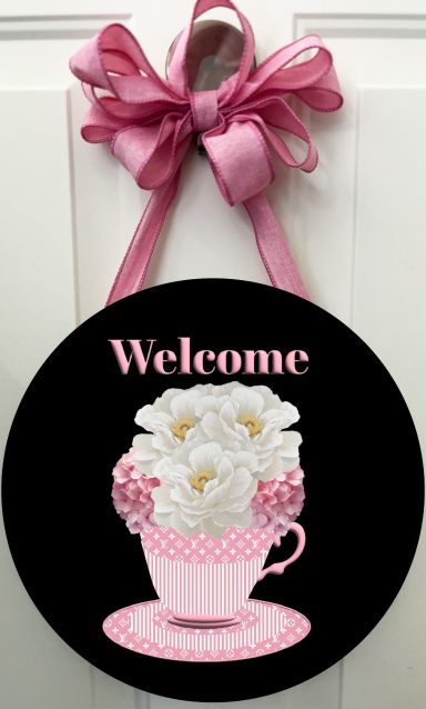Round sign featuring a pink teacup filled with white flowers and the word "Welcome."