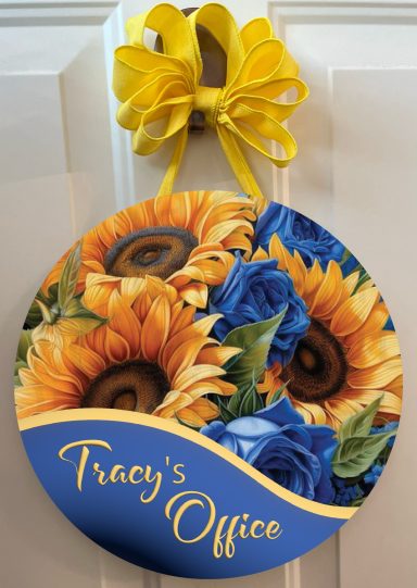 Round sign for "Tracy's Office" adorned with sunflowers and blue roses, tied with a yellow bow.