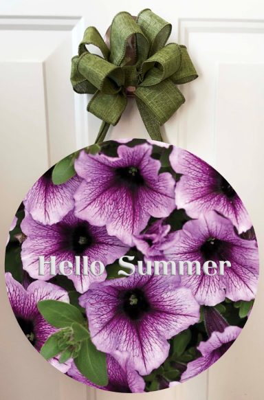 Round sign with purple petunia flowers and the text "Hello Summer" in white.